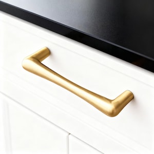 Op de afbeelding: Een goudkleurige kastgreep met een gebogen ontwerp, gemonteerd op een witte kastdeur. De greep heeft een gladde, matte afwerking en een moderne uitstraling. Het aanrechtblad boven de kast is zwart.
