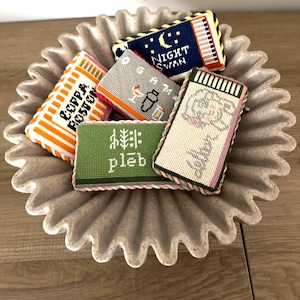 May include: A collection of colorful, hand-stitched needlepoint coasters with various designs and text, including "COPPA BOSTON", "NIGHT SWAN", "GAM", and "pleb". The coasters are arranged in a decorative, fluted, gray ceramic dish.