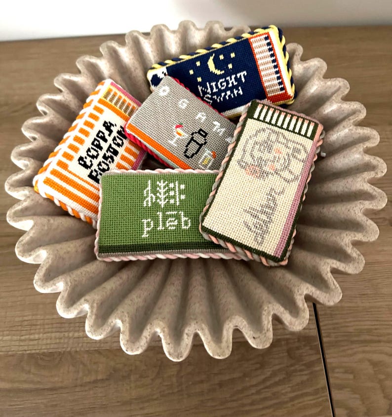 May include: A collection of colorful, hand-stitched needlepoint coasters with various designs and text, including "COPPA BOSTON", "NIGHT SWAN", "GAM", and "pleb". The coasters are arranged in a decorative, fluted, gray ceramic dish.