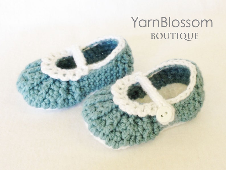 May include: A pair of crocheted baby shoes in a light blue color with white crocheted trim and a white button. The shoes have a simple, classic design.