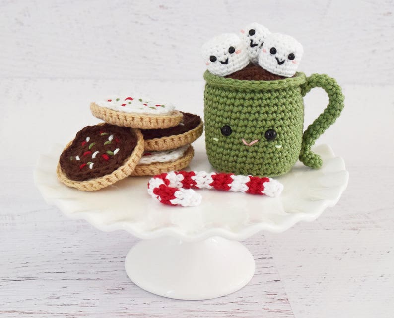 K&ouml;nnte beinhalten: Eine gr&uuml;ne geh&auml;kelte Tasse mit einem l&auml;chelnden Gesicht und wei&szlig;en geh&auml;kelten Marshmallows darauf. Die Tasse steht auf einem wei&szlig;en Kuchenst&auml;nder mit einem roten und wei&szlig;en Zuckerrohr und Schokoladenkeksen mit wei&szlig;en Streuseln.