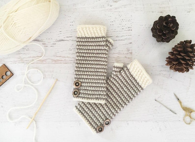 May include: A pair of beige and brown striped fingerless gloves with brown buttons. The gloves are made of crocheted yarn and are lying on a white wooden surface.