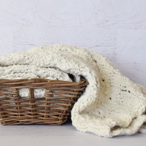 Può includere: Una coperta di uncinetto bianca piegata e posta in un cesto di vimini marrone.