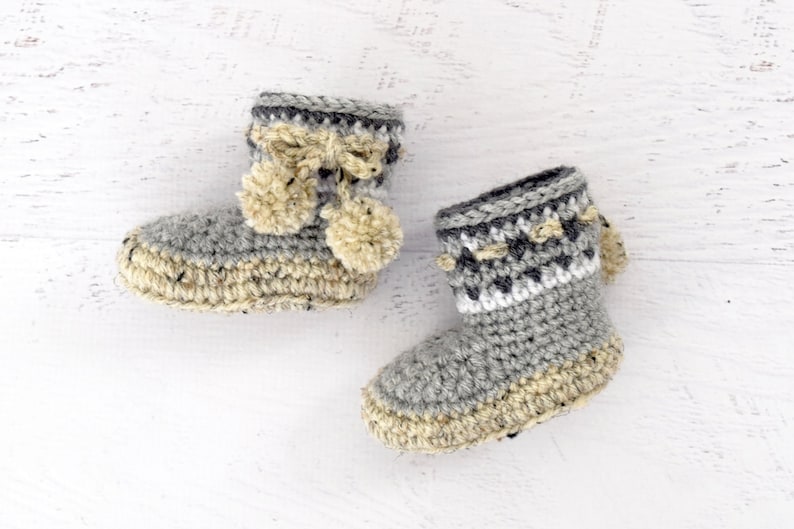 Pode incluir: Um par de sapatinhos de beb&eacute; em crochet cinzento e bege com pompons.