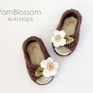 Op de afbeelding: Een paar bruine gehaakte babyschoentjes met witte bloemenversieringen en bruine bladeren. De schoentjes hebben een open teen en zijn gemaakt van zacht, comfortabel garen.