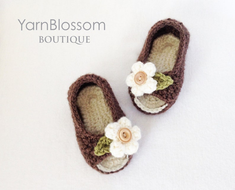Op de afbeelding: Een paar bruine gehaakte babyschoentjes met witte bloemenversieringen en bruine bladeren. De schoentjes hebben een open teen en zijn gemaakt van zacht, comfortabel garen.