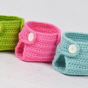 Op de afbeelding: Drie gehaakte babyluierhoezen in groen, roze en blauw met witte knopen.