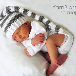 May include: A newborn baby wearing a white and grey striped crocheted hat with a long tail. The hat has a pointed tip and is made with a soft yarn. The baby is sleeping peacefully on a white blanket.