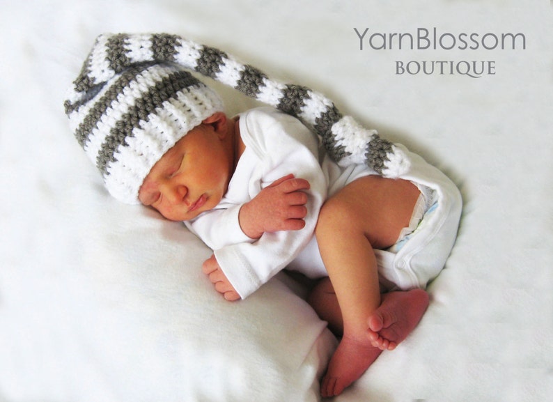 May include: A newborn baby wearing a white and grey striped crocheted hat with a long tail. The hat has a pointed tip and is made with a soft yarn. The baby is sleeping peacefully on a white blanket.