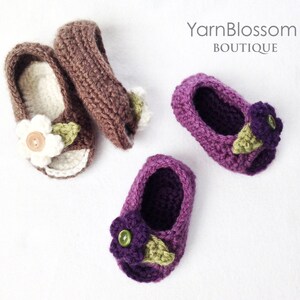 Op de afbeelding: Twee paar gehaakte babyschoentjes met bloemversieringen. Het ene paar is bruin met witte bloemen en het andere paar is paars met paarse bloemen. De schoentjes liggen op een witte achtergrond. YarnBlossom Boutique.