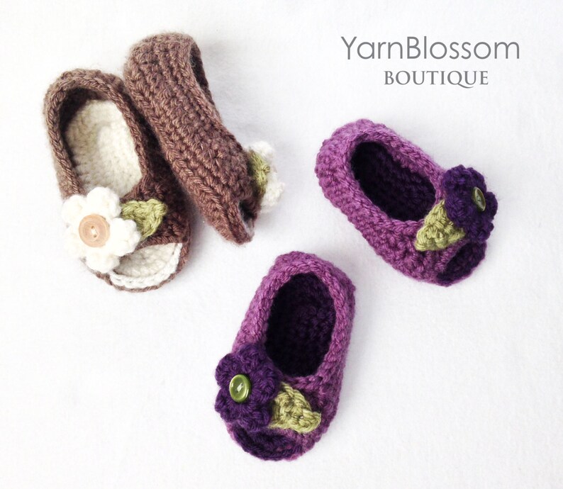 Op de afbeelding: Twee paar gehaakte babyschoentjes met bloemversieringen. Het ene paar is bruin met witte bloemen en het andere paar is paars met paarse bloemen. De schoentjes liggen op een witte achtergrond. YarnBlossom Boutique.