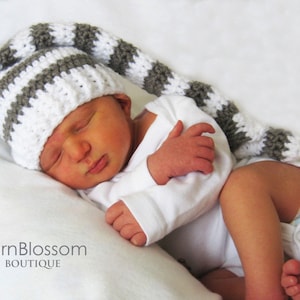 May include: A baby wearing a white and grey striped crocheted hat with a long tail. The hat has a pointed tip. The baby is wearing a white onesie and is sleeping.