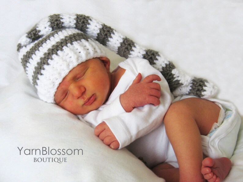 May include: A baby wearing a white and grey striped crocheted hat with a long tail. The hat has a pointed tip. The baby is wearing a white onesie and is sleeping.
