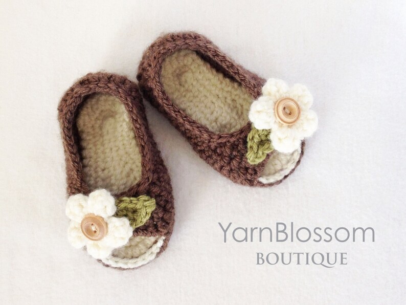 Op de afbeelding: Een paar bruine gehaakte babyschoentjes met witte bloemenversieringen en een knoop in het midden van elke bloem.