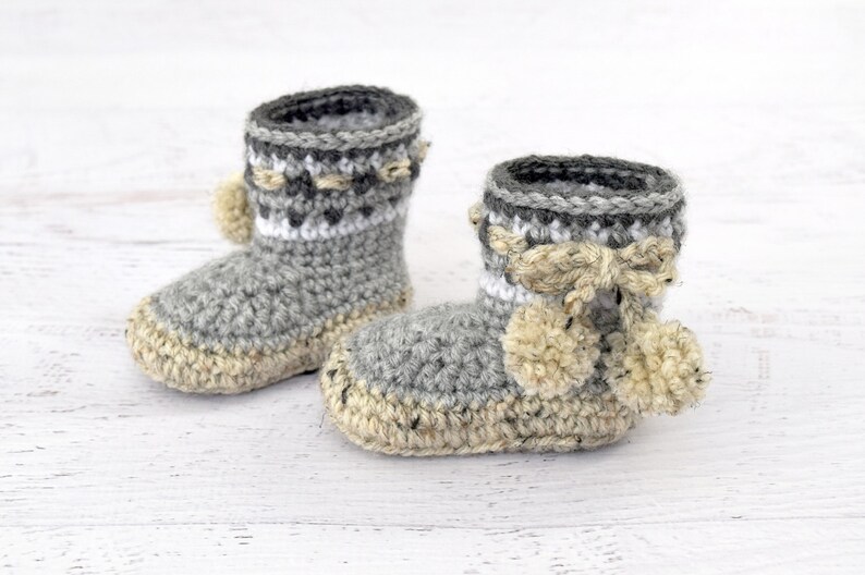 Pode incluir: Um par de sapatinhos de beb&eacute; em crochet cinzento e branco com pompons bege.