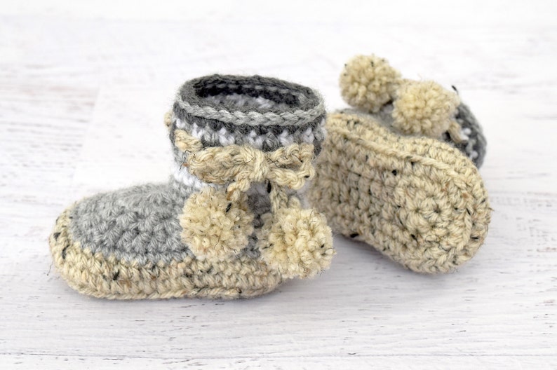Pode incluir: Um par de sapatinhos de beb&eacute; em crochet cinzento e bege com pompons.