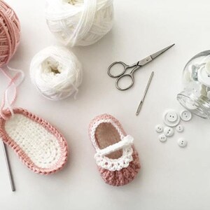 May include: A pair of pink and white crocheted baby shoes with white buttons and a crochet hook, yarn, and scissors on a white surface.
