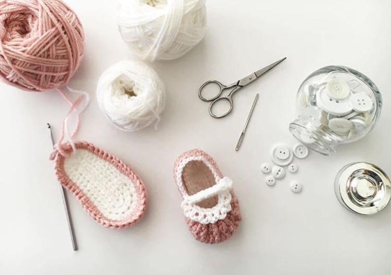 May include: A pair of pink and white crocheted baby shoes with white buttons and a crochet hook, yarn, and scissors on a white surface.