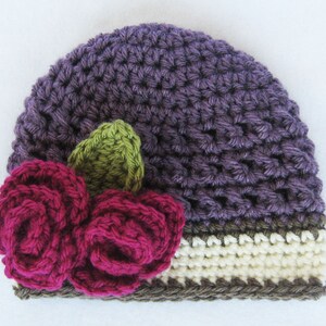May include: Purple crocheted hat with a brown and white crocheted band and a pink and green flower embellishment.