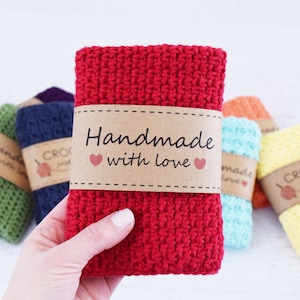 Könnte beinhalten: Eine Hand hält einen roten gehäkelten Waschlappen mit einem braunen Papierband mit der Aufschrift "Handmade with love" und zwei rosa Herzen. Weitere farbenfrohe gehäkelte Waschlappen in Grün, Blau, Orange und Gelb sind im Hintergrund.