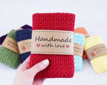 Puede incluir: Una mano sostiene un paño de cocina rojo tejido a ganchillo con una banda de papel marrón que dice "Handmade with love" y dos corazones rosas. Otros paños de cocina tejidos a ganchillo de colores en verde, azul, naranja y amarillo están en el fondo.