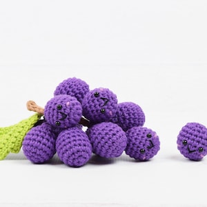 Pode incluir: Um cacho de uvas de crochê roxo com carinhas felizes. As uvas estão dispostas em um cacho com um caule de crochê verde.
