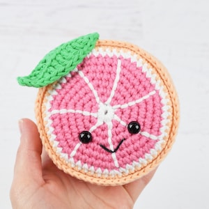 Può includere: Un pompelmo all'uncinetto rosa e bianco con una foglia verde e occhi neri. Il pompelmo ha un viso sorridente.