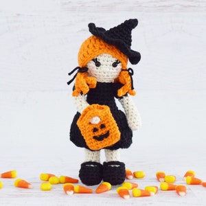 May include: A crocheted witch doll wearing a black hat and orange dress, holding a pumpkin-shaped trick-or-treat bag. The doll is surrounded by orange and white candy corn.