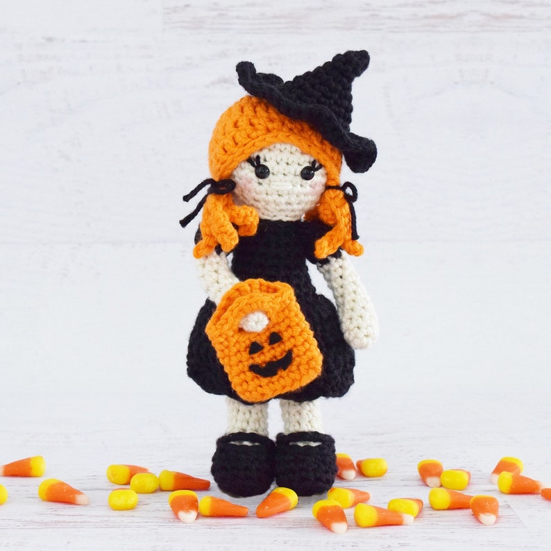 May include: A crocheted witch doll wearing a black hat and orange dress, holding a pumpkin-shaped trick-or-treat bag. The doll is surrounded by orange and white candy corn.