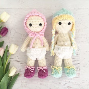 Puede incluir: Dos muñecas de crochet que llevan sombreros y pañales. La muñeca de la izquierda lleva un sombrero rosa y la muñeca de la derecha lleva un sombrero azul claro y amarillo.