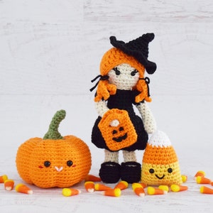 May include: A crocheted witch doll with orange hair, a black hat, and a black dress holding a pumpkin-shaped trick-or-treat bag. The doll is standing next to a crocheted orange pumpkin and a crocheted candy corn.