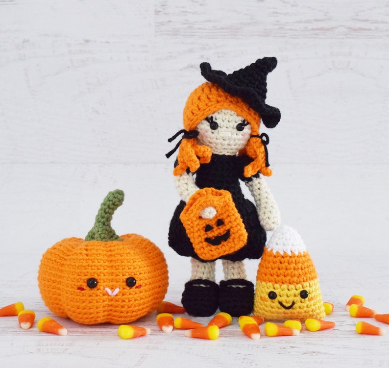 May include: A crocheted witch doll with orange hair, a black hat, and a black dress holding a pumpkin-shaped trick-or-treat bag. The doll is standing next to a crocheted orange pumpkin and a crocheted candy corn.
