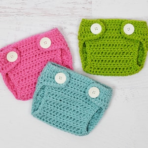Op de afbeelding: Drie gehaakte babyluierhoezen in roze, groen en blauw met witte knopen.