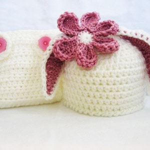 May include: A white crocheted baby hat with pink bunny ears and a pink flower on top. The hat is made with a soft yarn and is perfect for a newborn baby.