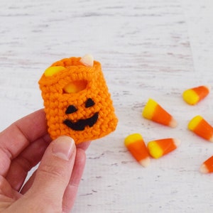 May include: A small orange crocheted pumpkin-shaped bag with a black jack-o'-lantern face. The bag is filled with candy corn.