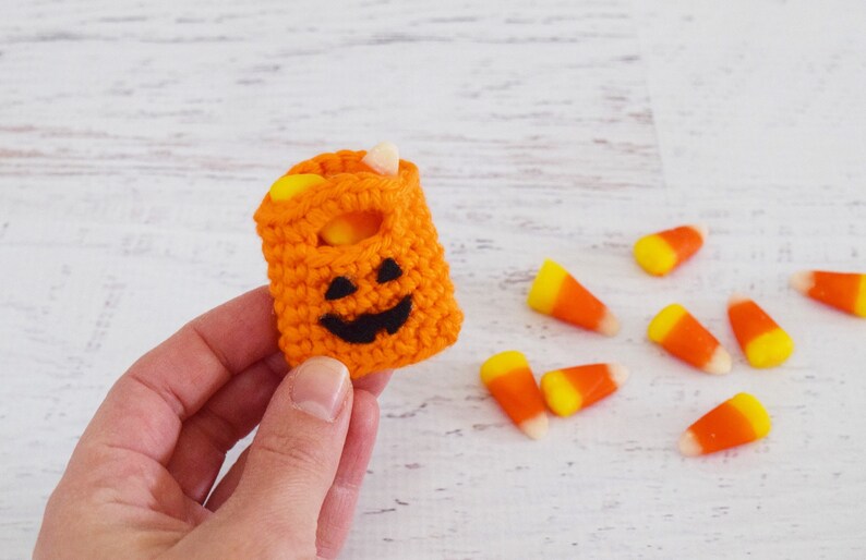 May include: A small orange crocheted pumpkin-shaped bag with a black jack-o'-lantern face. The bag is filled with candy corn.
