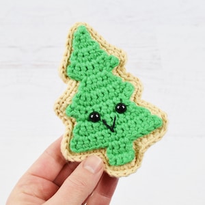 May include: A green crocheted Christmas tree with a smiling face and black eyes. The tree is surrounded by a tan crocheted border.