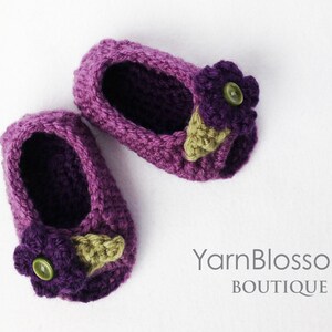 Op de afbeelding: Een paar paarse gehaakte babyschoentjes met een paarse bloem en een groen blaadje als versiering op elke schoen. YarnBlossom Boutique.