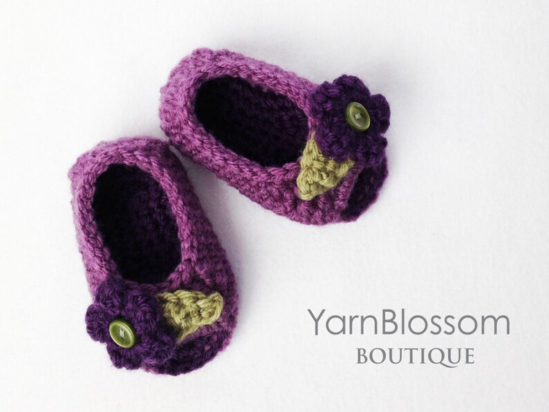 Op de afbeelding: Een paar paarse gehaakte babyschoentjes met een paarse bloem en een groen blaadje als versiering op elke schoen. YarnBlossom Boutique.