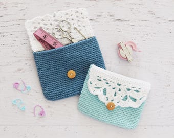 Può includere: Due pochette all'uncinetto con bordo in pizzo bianco e bottoni in legno. Una pochette è blu e l'altra è azzurro chiaro. La pochette blu contiene un metro a nastro rosa e delle forbici.