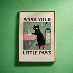 Op de afbeelding: Een ingelijste kunstdruk met een zwarte kat die zijn poten wast in een roze wastafel. De print bevat de tekst "WASH YOUR LITTLE PAWS". De achtergrond toont een groene betegelde muur en een handdoek met een bloemenpatroon.