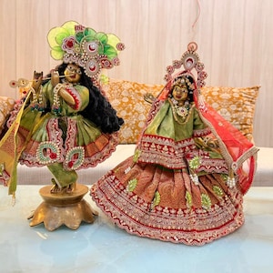 May include: Two decorative statues of deities, one playing a flute, the other with a veil. Both are dressed in green and red embroidered garments, adorned with jewels and elaborate headpieces. They are placed on a table.