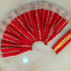 Puede incluir: Un abanico de terciopelo rojo con adornos y adornos plateados. El abanico tiene una bolsa roja a juego con detalles dorados y un cordón dorado. El abanico está abierto.