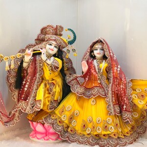 May include: Two statues of deities, one playing a flute, the other with a hand raised. Both are dressed in yellow and red garments with detailed gold embroidery. The figures have detailed facial features and are set against a neutral background.