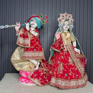 May include: Two ornate statues of deities. One figure, in red and gold attire, plays a flute. The other figure, in a similar style, wears a crown and holds a hand up. Both statues are on a white base.