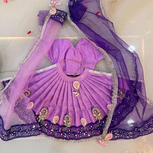 Könnte beinhalten: Ein traditionelles indisches Outfit in Lilatönen. Das Ensemble umfasst einen plissierten Rock, ein kurzärmeliges Oberteil und einen transparenten Schal mit aufwendigen Stickereien und Verzierungen. Das Outfit ist mit dekorativen Blumen- und Perlenakzenten verziert.