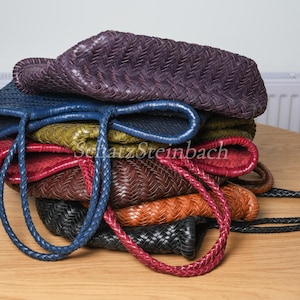 May include: A collection of woven leather tote bags in shades of purple, blue, green, red, brown, orange, and black. Each bag features braided handles. The bags are stacked on a wooden surface.