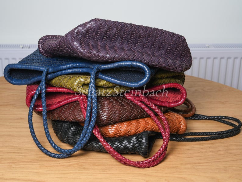 May include: A collection of woven leather tote bags in shades of purple, blue, green, red, brown, orange, and black. Each bag features braided handles. The bags are stacked on a wooden surface.