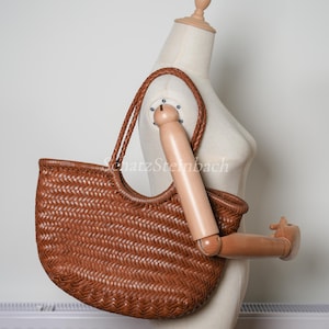 May include: A brown woven tote bag with a braided handle and a curved top. The bag is displayed on a mannequin. The bag's woven pattern is visible, and the colour is a warm brown.
