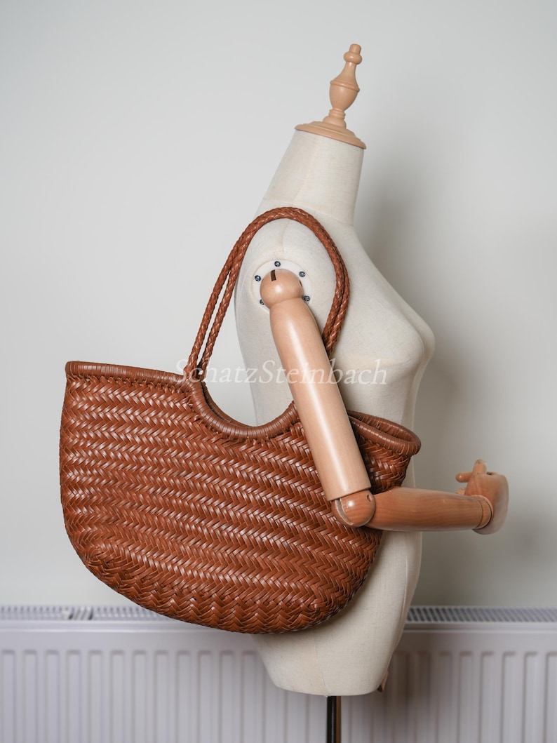 May include: A brown woven tote bag with a braided handle and a curved top. The bag is displayed on a mannequin. The bag's woven pattern is visible, and the colour is a warm brown.